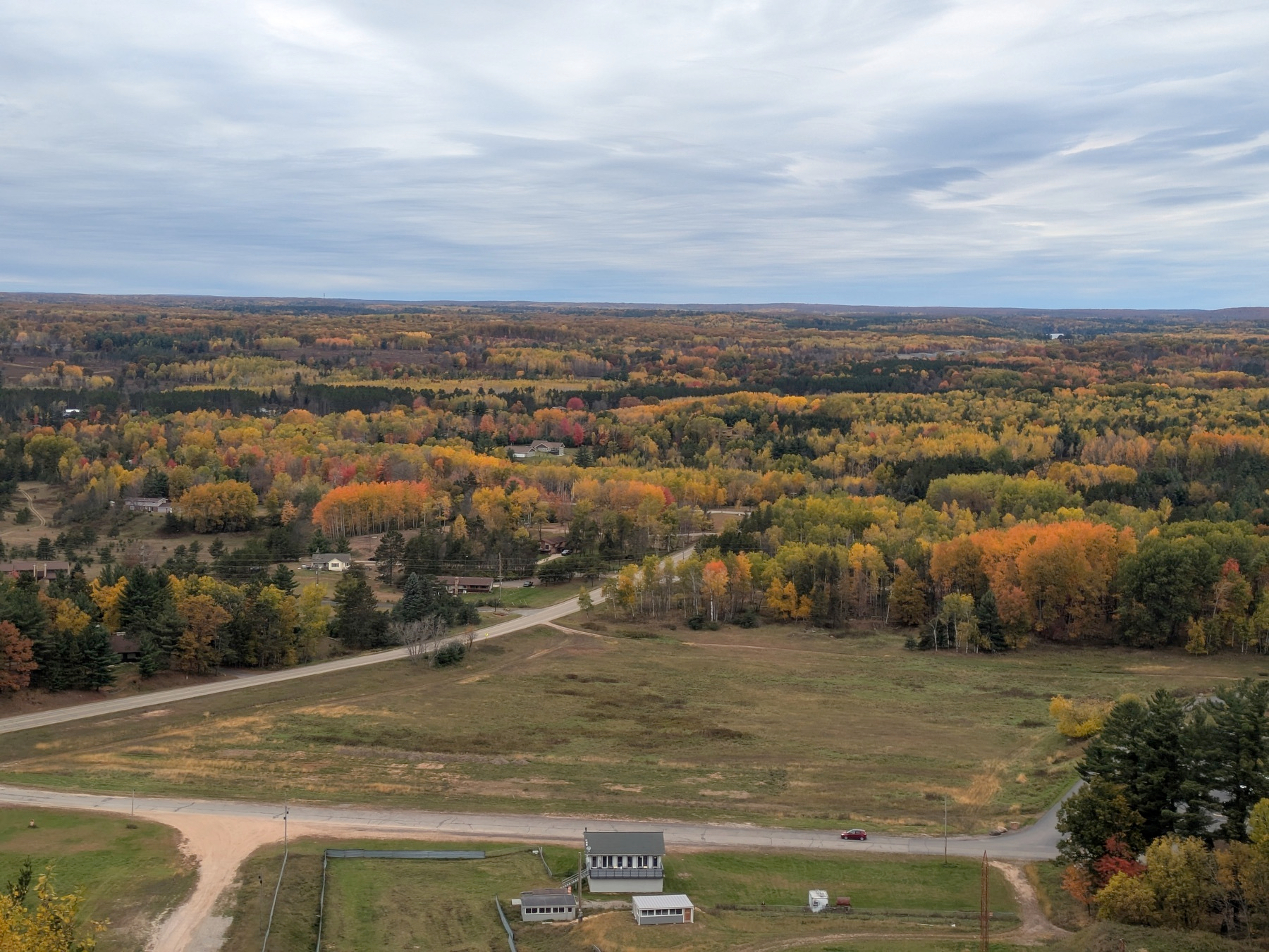 Auto-generated description: A vast landscape features autumn foliage with vibrant hues of orange, yellow, and green, seen from an elevated viewpoint with a small building in the foreground.