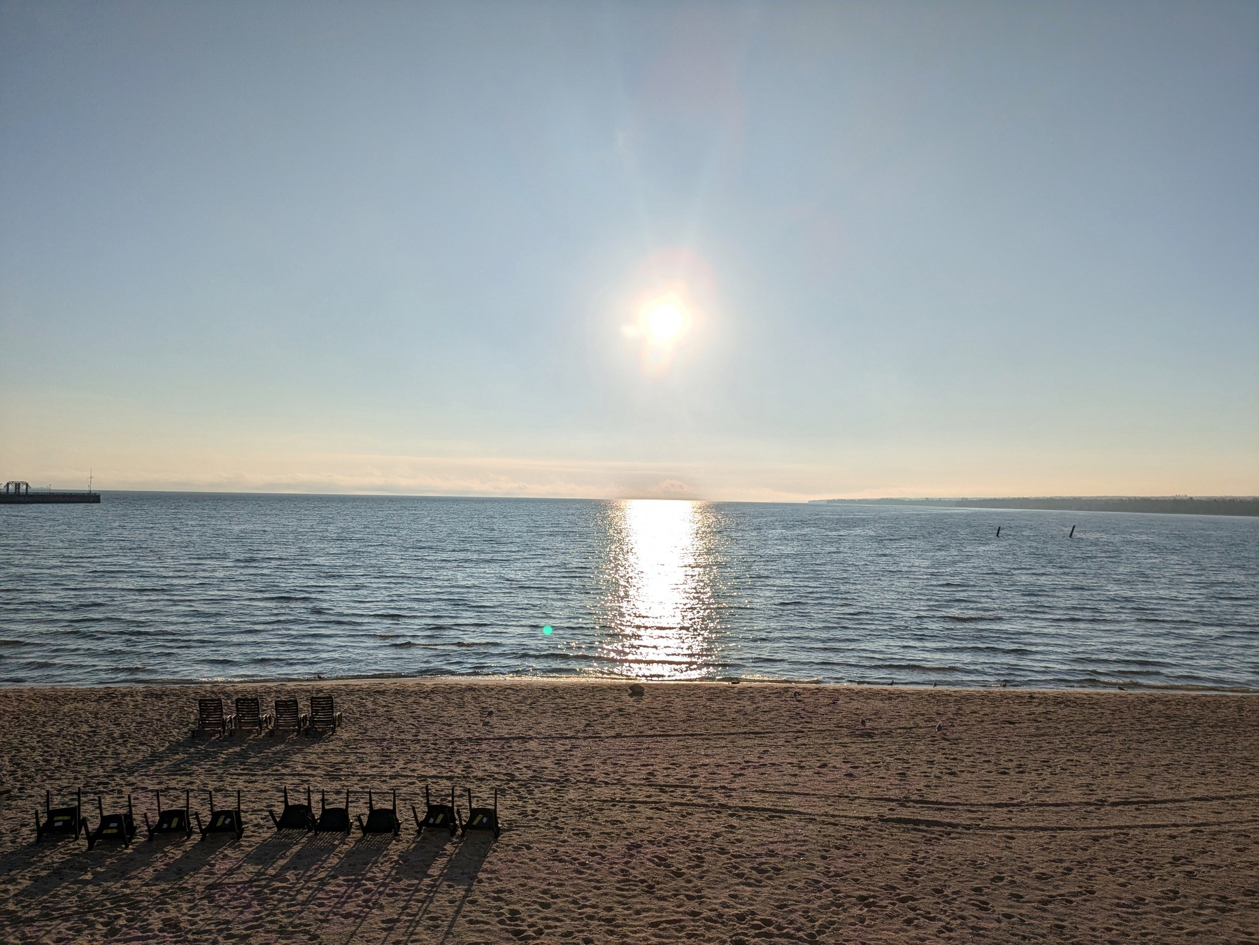 Auto-generated description: A serene beach scene features the sun setting over the ocean, casting reflections on the water, with empty lounge chairs arranged on the sandy shore.
